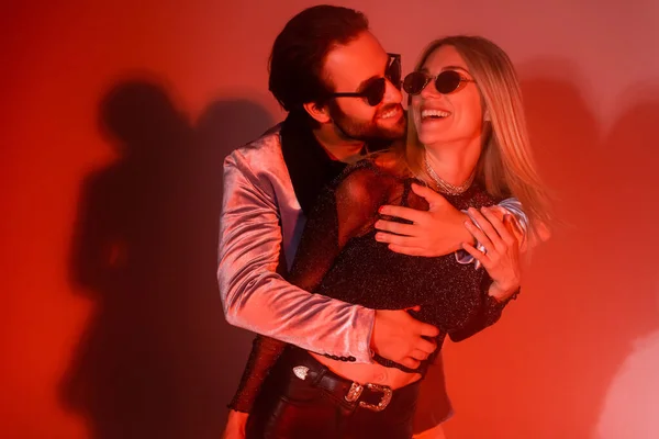 Smiling man embracing blonde girlfriend in sunglasses during party on red background - foto de stock