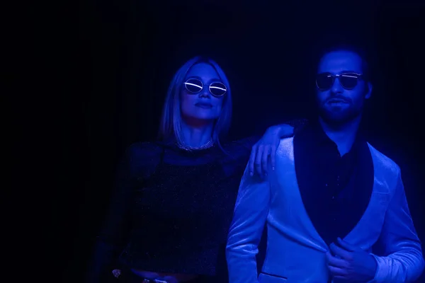 Fashionable couple in sunglasses standing isolated on black with blue lighting — Stock Photo