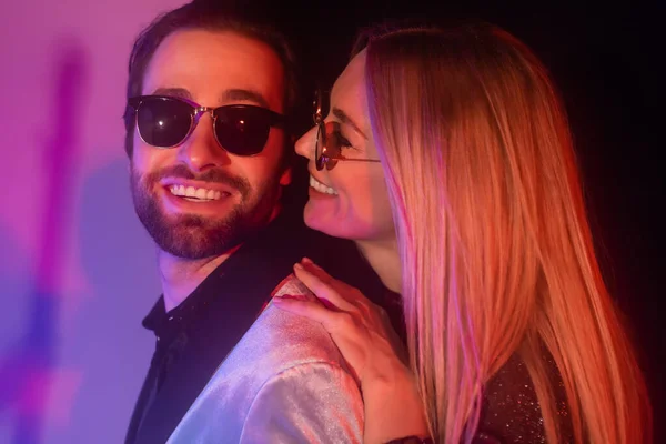 Cheerful blonde woman in sunglasses hugging boyfriend in jacket on purple background - foto de stock