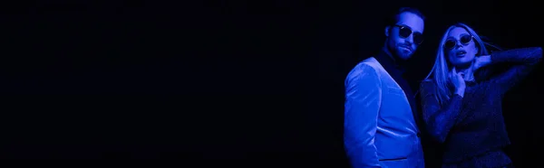 Fashionable man in sunglasses standing near girlfriend in sunglasses isolated on black with blue lighting, banner — Fotografia de Stock