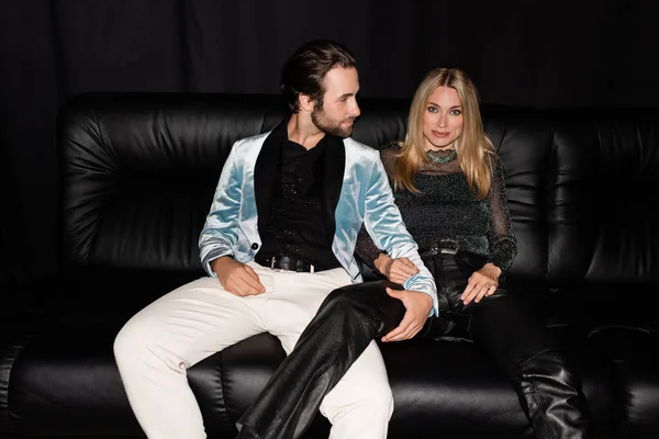 Stylish man touching blonde girlfriend on leather couch isolated on black — Photo de stock