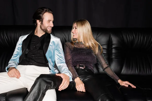 Веселая стильная пара, смотрящая друг на друга, сидя на кожаном диване, изолированном на черном — стоковое фото