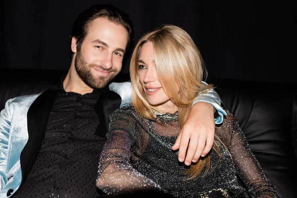 Smiling man hugging stylish girlfriend on leather couch isolated on black — Fotografia de Stock