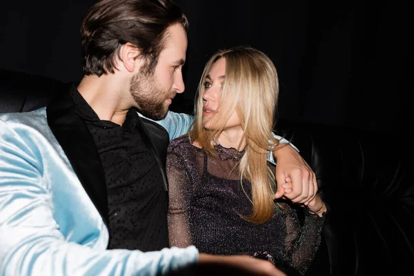 Stylish man hugging blonde girlfriend on leather couch during party isolated on black — Fotografia de Stock