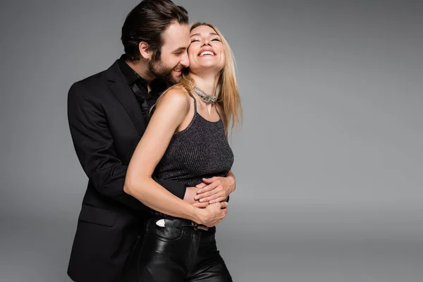 Bearded man in jacket hugging smiling blonde girlfriend isolated on grey - foto de stock