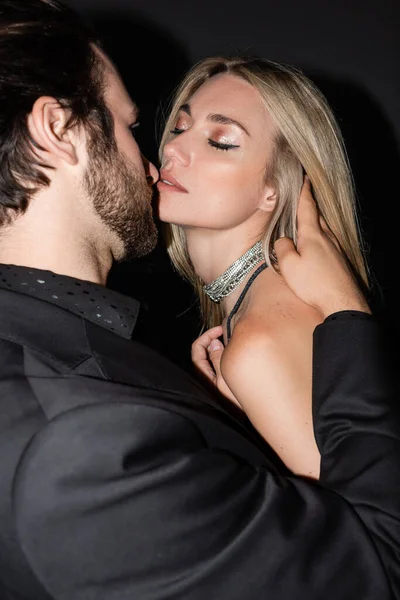 Sexy blonde woman kissing bearded boyfriend in jacket isolated on black — Stock Photo