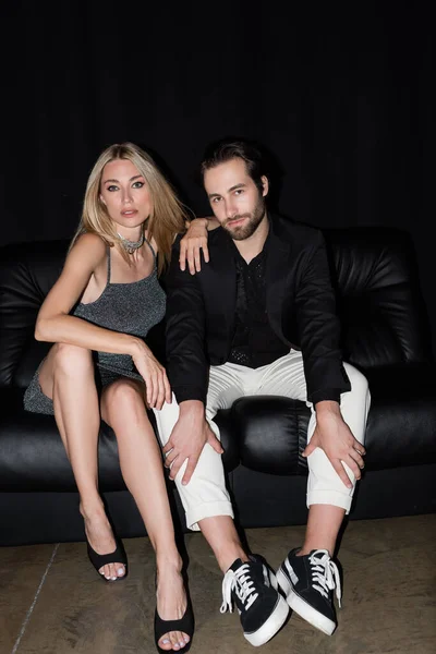 Blonde woman in dress looking at camera near boyfriend on leather couch on black background — Fotografia de Stock