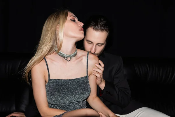 Young man kissing shoulder of sexy blonde girlfriend on leather couch isolated on black — Photo de stock