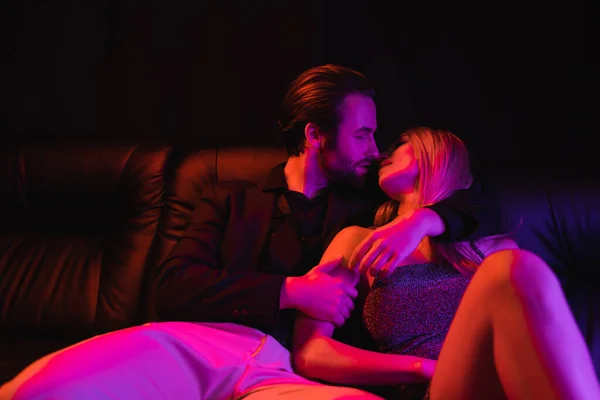 Bearded man kissing blonde girlfriend on leather couch with lighting isolated on black — Photo de stock