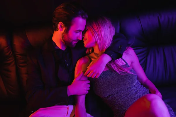 Side view of young man kissing sexy girlfriend on leather couch with lighting isolated on black — Photo de stock