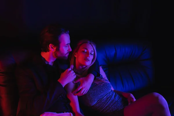 Passionate man hugging girlfriend in dress on leather couch with lighting isolated on black — Photo de stock