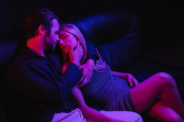 Sexy man touching and kissing blonde girlfriend on leather couch with lighting isolated on black — стоковое фото