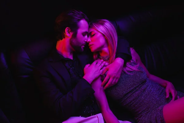 Side view of sexy couple kissing on leather couch with lighting during party isolated on black — Stock Photo