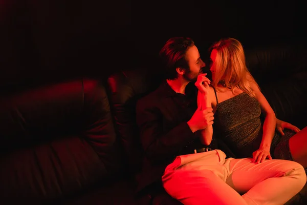 Woman in dress touching young boyfriend on leather couch with lighting isolated on black — Photo de stock