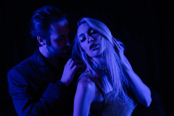 Man in jacket touching sexy girlfriend with blue lighting isolated on black — Stock Photo