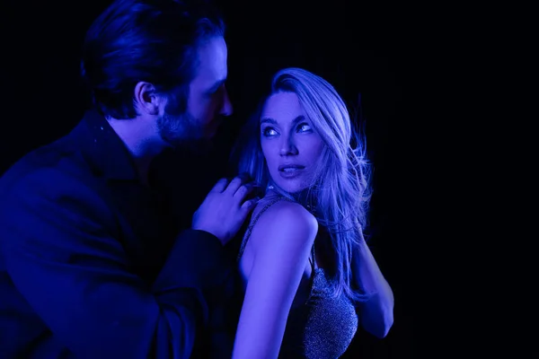 Young man touching seductive blonde girlfriend with blue lighting isolated on black — Photo de stock