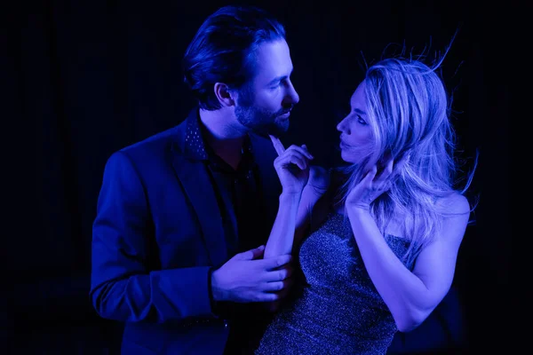 Side view of sexy woman in dress touching boyfriend in jacket with blue lighting isolated on black — Photo de stock