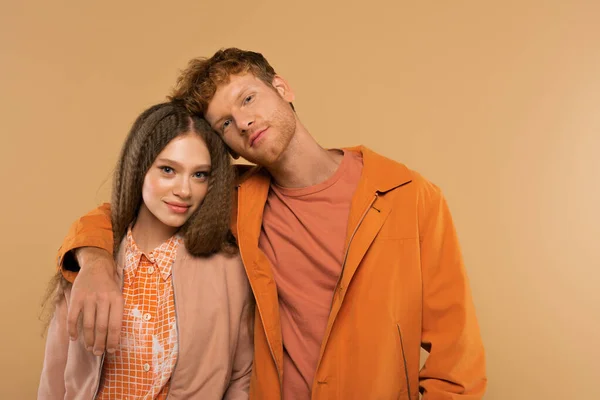 Redhead young man in orange jacket hugging cheerful girlfriend isolated on beige — стоковое фото