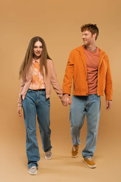 Full length of happy young couple in autumnal outfits and gumshoes holding hands while walking on beige - foto de stock