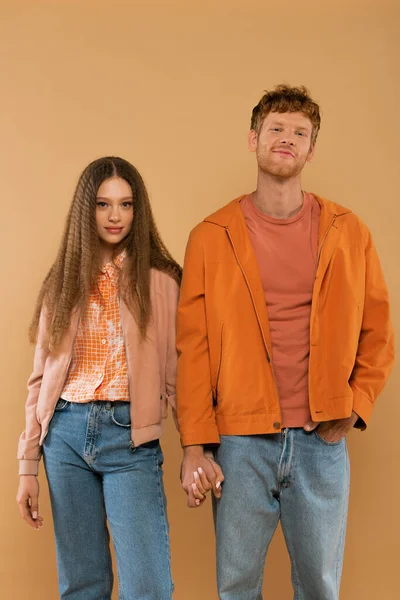 Happy young couple in autumnal outfits holding hands while standing isolated on beige — Fotografia de Stock