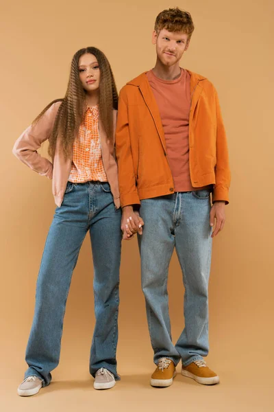 Full length of young couple in autumnal outfits and gumshoes holding hands while standing on beige - foto de stock