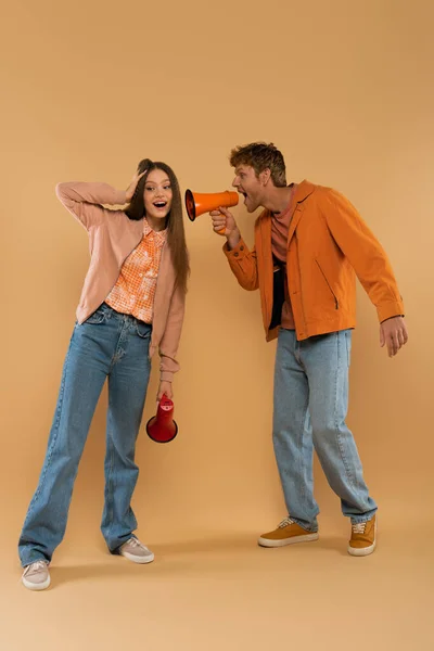 Full length of redhead man screaming in loudspeaker near amazed girlfriend on beige — Foto stock