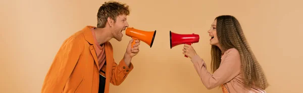 Side view of young couple screaming in loudspeakers and smiling isolated on beige, banner — Stockfoto