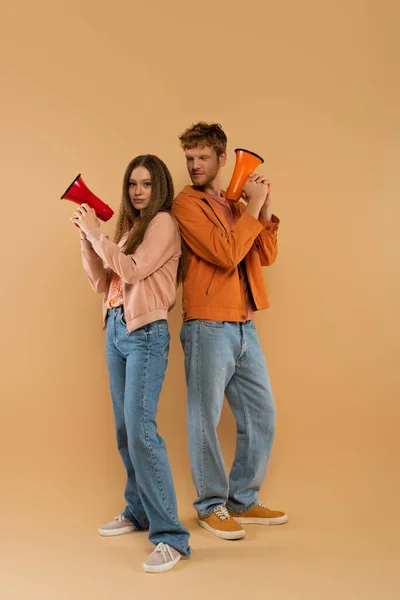 Full length of young couple holding loudspeakers and standing on beige - foto de stock
