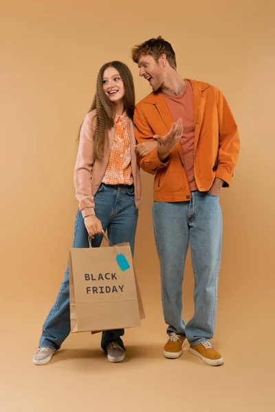 Повнометражний молодий чоловік розмовляє біля щасливої дівчини тримає сумку з чорною п'ятничною пляшкою на бежевому — стокове фото