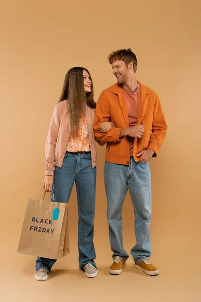 Full length of young man looking at happy girlfriend holding paper bag with black friday lettering on beige — Photo de stock