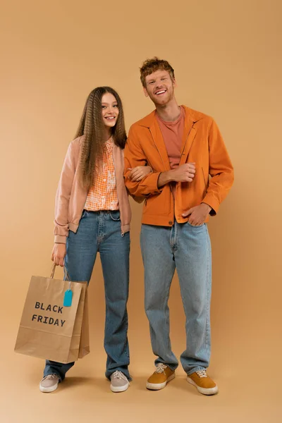 Full length of young man posing near happy girlfriend holding paper bag with black friday lettering on beige - foto de stock