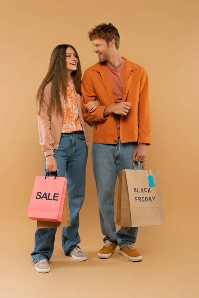 Full length of young couple holding shopping bags with sale and black friday lettering on beige — стоковое фото