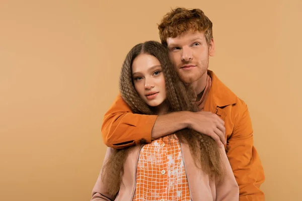 Young redhead man hugging girlfriend with wavy hair isolated on beige — стоковое фото