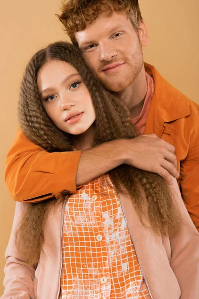 Smiling young redhead man hugging girlfriend with wavy hair isolated on beige — стоковое фото