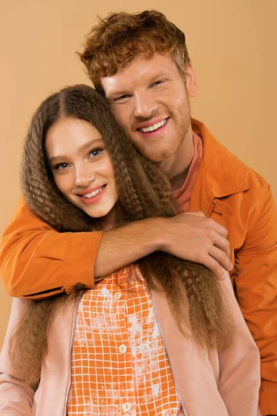 Cheerful young redhead man hugging happy girlfriend with wavy hair isolated on beige — стоковое фото