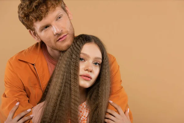 Young man with red hair hugging pretty girlfriend looking at camera isolated on beige — Fotografia de Stock