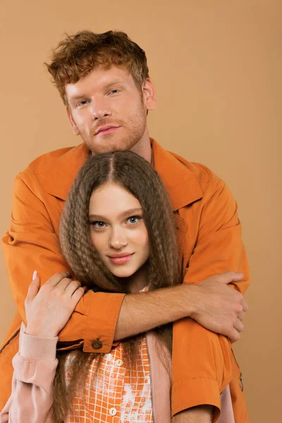 Young man with red hair looking at camera and hugging pretty girlfriend isolated on beige — Stock Photo