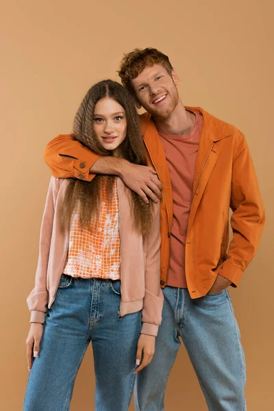 Young cheerful man with red hair looking at camera and hugging pretty girlfriend isolated on beige — стоковое фото