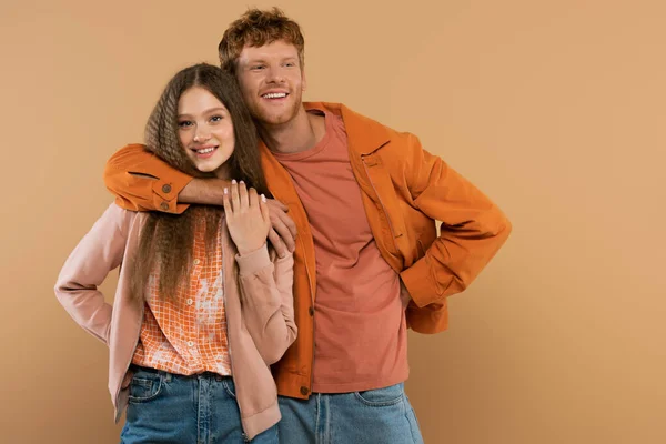 Young happy man with red hair looking at camera and hugging pretty girlfriend isolated on beige — Stockfoto