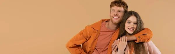 Young happy man with red hair looking at camera and hugging pretty girlfriend isolated on beige, banner — Stockfoto