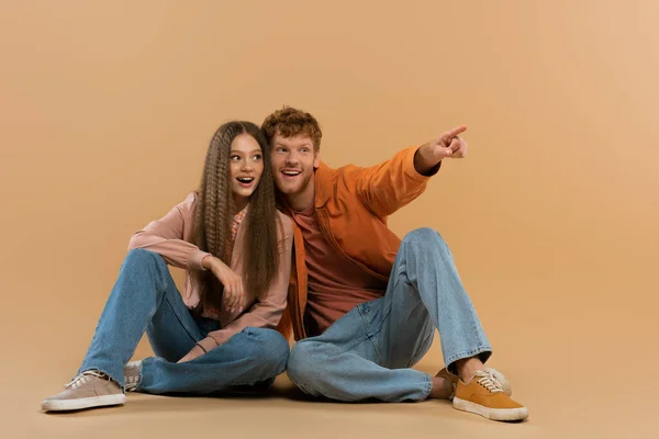 Happy and young redhead man pointing with finger while sitting near girlfriend on beige — Fotografia de Stock