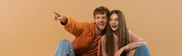 Happy and young redhead man pointing with finger while sitting near girlfriend isolated on beige, banner - foto de stock