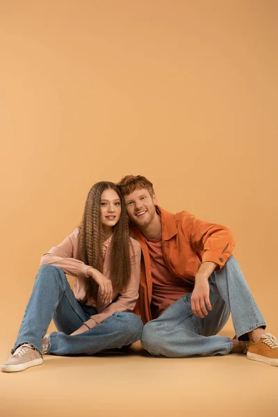 Full length of cheerful young couple in jeans and jackets sitting and smiling on beige — Stock Photo