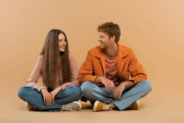 Full length of cheerful young couple in jeans and jackets sitting and looking at each other on beige — Stock Photo