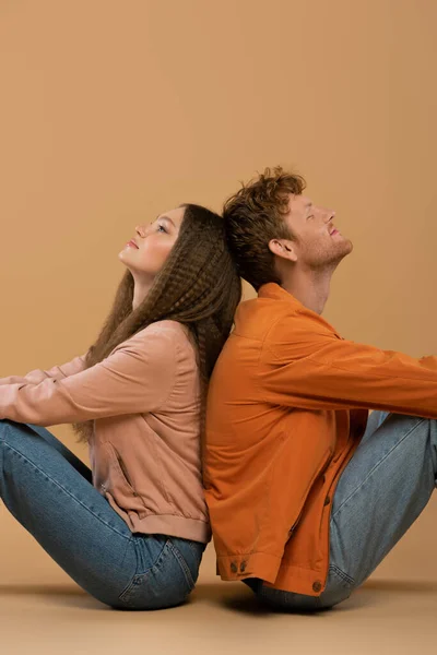 Side view of smiling young man with red hair sitting back to back with girlfriend on beige — Photo de stock