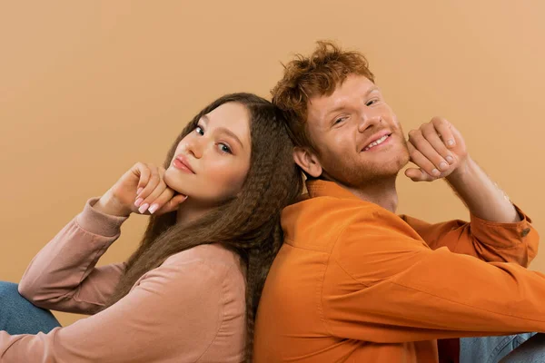 Joyful young man with red hair sitting back to back with pretty girlfriend isolated on beige - foto de stock