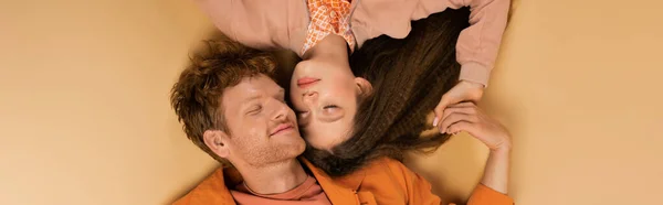 Top view of happy and young redhead man lying together with pretty girlfriend on beige, banner — стоковое фото