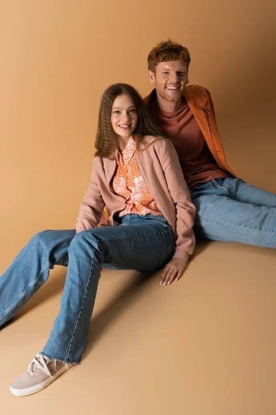 Full length of cheerful and young couple in denim jeans sitting on beige — стоковое фото