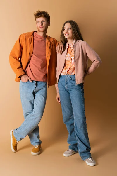 Full length of cheerful and young couple in denim jeans standing on beige — стоковое фото