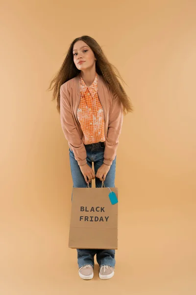 Teenage girl holding paper bag with black friday lettering while standing on beige — стоковое фото